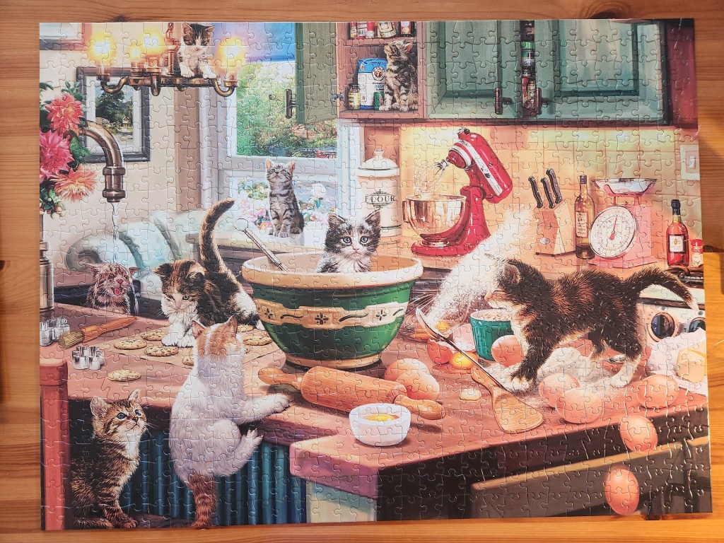 Completed jigsaw puzzle showing kittens in the kitchen causing mischief. One is in a cupboard looking up at a falling container. One sits on the chandelier looking down. One sits in a mixing bowl. One is stepping on a spoon of flour throwing it in their face. Another is on a parchment paper sheet of cookies playing with them. Another is sitting under the running faucet in the sink. A few others sit around watching the activity amongst eggs, rolling pins, bowls, and other assorted baking supplies.