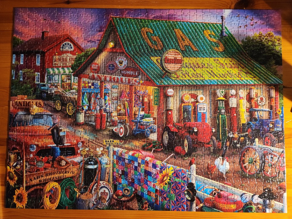 Completed jigsaw puzzle showing a busy country scene with a gas station, antique show, and general store. The image also shows a red tractor and a blue tractor, as well as a colorful quilt. There are some roosters and chickens and cars as well.